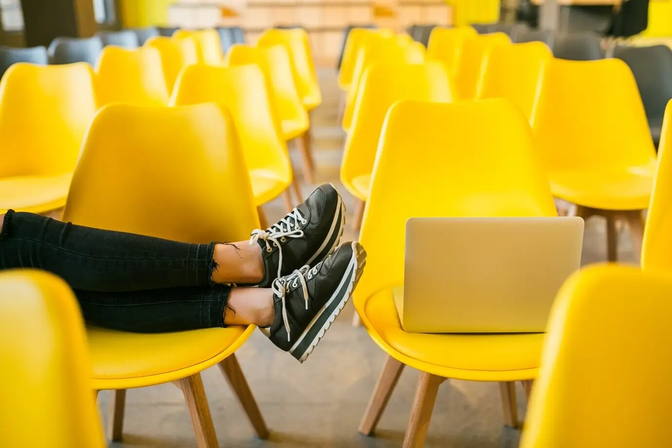 Close-up of the legs of a young stylish woman seated in a lecture hall with a laptop, in a classroom with many yellow chairs; sneakers, a footwear fashion trend.