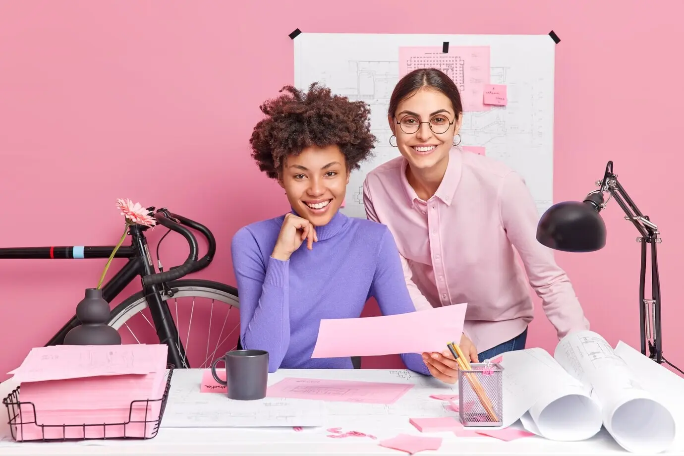 Two diverse, creative women collaborate on blueprints for a new project, posing in a coworking space and enjoying their occupation.